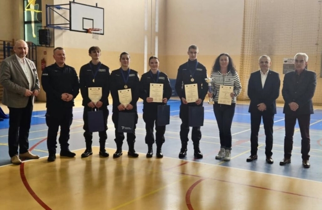Dziewięć osób stoi w sali gimnastycznej. 4 zawodników konkursu, uczniów klas policyjnych w mundurach trzyma w ręku dyplomy, na szyjach mają medale, a ich opiekun, stojący obok nich po prawej stornie trzyma w prawej ręce puchar, a w lewej dyplom. Pozostałe cztery osoby są ubrane w stroje cywilne i stoją po bokach grupy w mundurach. W tle widać kosz do koszykówki oraz linie namalowane na podłodze hali.