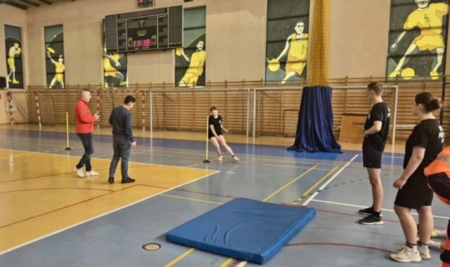 Pięć osób w hali sportowej. Trzech zawodników pokonuje tor przeszkód, podczas gdy dwóch sędziów obserwuje ich z boku. W tle widoczne są bramki oraz drabinki sportowe. Na podłodze znajduje się materac zapewniający bezpieczeństwo.