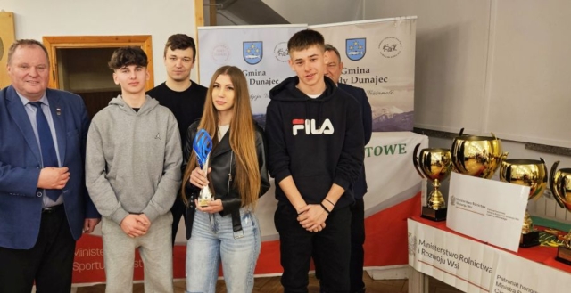 rajd_podhalanski04 Zdjęcie przedstawia młodzież - uczestników rajdu oraz 3 osoby dorosłe. Jedna uczennica trzyma w ręku trofeum sportowe. W tle banery reklamowe Gminy Biały Dunajec, po prawej stronie stolik z pucharami i informacja o patronacie honorowym wydarzenia.