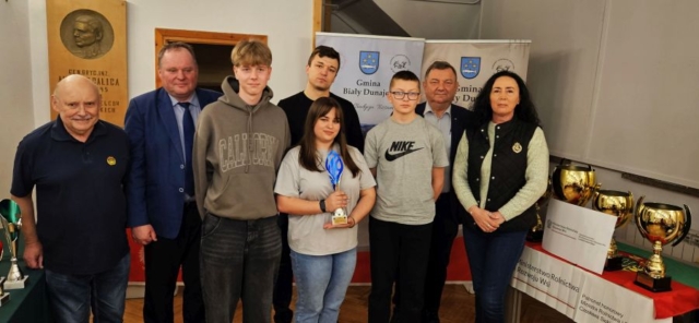 rajd_podhalanski05 Zdjęcie przedstawia uczestników rajdu wraz z opiekunem oraz pracowników Urzędu Gminy. Jedna uczennica trzyma w ręku trofeum sportowe. W tle banery reklamowe, tablica pamiątkowa, po prawej stronie stoli z pucharami oraz informacją o patronacie wydarzenia.