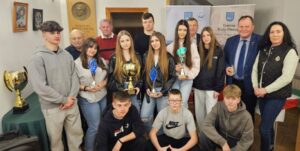 Zdjęci przedstawia grupę młodzieży wraz z opiekunem i organizatorami rajdu pozujących do zdjęcia. 4 osoby trzymają w rękach puchary. W tle banery reklamowe gminy Biały Dunajec o raz tablica pamiątkowa. 