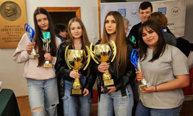 rajd_podhalanski09 Zdjęcie przedstawia 4 uczestniczki radju, trzymające w rękach puchary i trofea sportowe. W tle osoby dorosłe, tablica pamiątkowa poświęcona twórcy pułków strzeleckich podhalańskich oraz fragmenty banerów Urzędu Gminy.