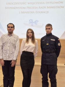 Zdjęcie przedstawia trzech Stypendystów Szkoły. Dwóch uczniów i jedną uczennicę, ubranych na galowo, jeden z uczniów w mundurze klasy policyjnej. Uczniowie stoją w auli, w tle ekran z napisem wielkimi literami  uroczystość wręczenia dyplomów stypendium prezesa rady ministrów i ministra edukacji. 