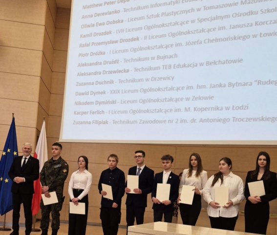 Zdjęci przedstawia 8 uczniów w strojach galowych, w tym jeden uczeń w mundurze klasy wojskowej oraz przedstawiciela komisji wręczającej dyplomy, na uroczystości. Uczniowie stoją w auli, w tle ekran z nazwiskami Stypendystów. Po prawej stronie flagi Polski oraz Unii Europejskiej.