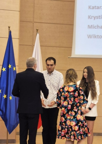 Zdjęcie przedstawia 2 uczniów w strojach galowych i dwóch przedstawicieli kuratorium, wręczających dyplomy za wyniki w nauce. Z lewej strony flagi Polski i Unii Europejskiej.