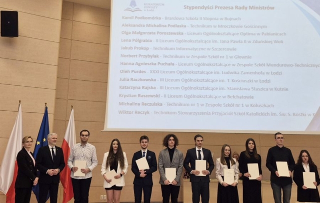 Zdjęcie przedstawia uczniów w strojach galowych stojących w auli. Są tez przedstawiciele kuratorium. Po lewej stronie flagi Polski oraz Unii Europejskiej. W tle ekran z nazwiskami Stypendystów wraz z nazwami szkół, do których uczęszczają.