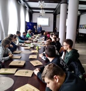 Zdjęcie przedstawia salę konferencyjną, przy stole siedzą uczestnicy szkolenia. W tle ekran z wyświetlaną prezentacją, a także banery koloru granatowego. Na stole teczki z materiałami szkoleniowymi. 