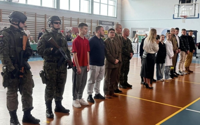 Zdjęcie przedstawia uczniów i p. dyrektor, przemawiającą przez mikrofon i opiekuna stojących w rzędzie na sali gimnastycznej. W tle drabinki oraz stoiska szkól, prezentujących swoją ofertę edukacyjną.