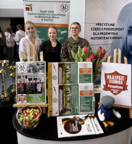 Zdjęci przestawia uczennice reprezentujące szkołę na targach edukacyjnych oraz stoisko szkolne. w tle rollupy Zespołu Szkół Centrum Kształcenia Zawodowego im. Wincentego Witosa w Bujnach oraz anton-haering.