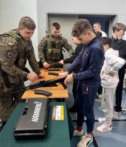 Zdjęcie przedstawia uczniów reprezentujących szkołę oraz uczniów biorących udział w targach edukacyjnych podczas prezentacji i składania broni.
