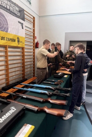 Zdjęcie przedstawia stoisko szkoły, uczniów reprezentujących szkołę podczas targów edukacyjnych, a także zainteresowanych uczniów, którzy biorą udział w prezentacji i składaniu broni.