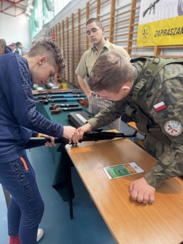 Zdjęcie przedstawia uczniów reprezentujących szkołę oraz uczestnika na targach edukacyjnych. W tle drabinki szkolne i część baneru reklamującego strzelnicę pneumatyczną w ZS CKZ Bujny.