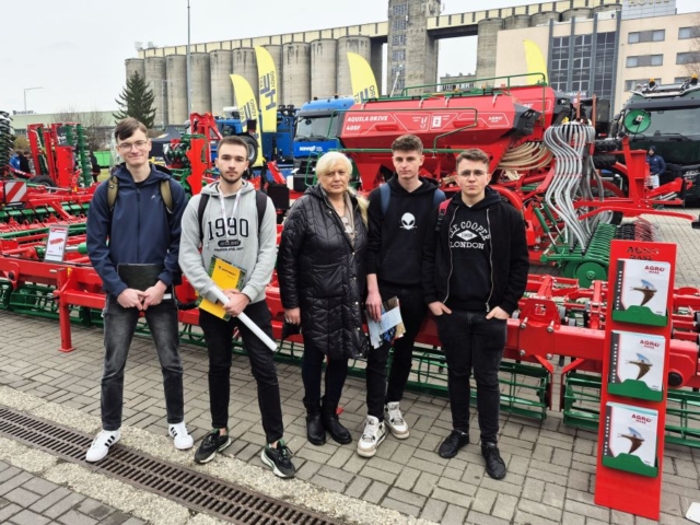 targi_kielce2 Zdjęcie przedstawia 4 uczestników targów wraz z opiekunem stojących na tle maszyny rolniczej w kolorze czerwonym. W tle budynki i inne maszyny.