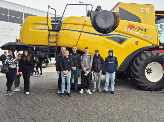 targi_kielce6 Zdjęcie przedstawia 9 uczestników targów, stojących na zewnątrz na tle maszyny rolniczej koloru żółtego. Na maszynie napis modelu oraz napis New Holland, Agros.