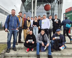 Zdjęcie przedstawia uczestników wycieczki na targi rolnicze wraz z opiekunem. Osoby na zdjęciu stoją lub siedzą na schodach przed budynkiem targów. 
