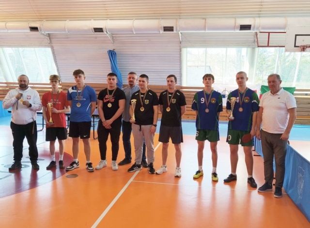 7 zawodników stoi w hali sportowej. Wszyscy mają na szyjach medale, 3 osoby trzymają w rękach puchary. Widoczni także 3 opiekunowie drużyn. Jeden z zawodników trzyma też paletkę tenisową.