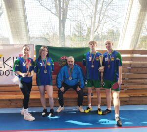 2 zawodniczki i dwóch zawodników w strojach sportowych koloru zielonego, granatowego i czarnego stoją w hali sportowej na tle dużego okna przesłoniętego siatką. Na szyjach mają medale. Dwie osoby trzymają w rękach puchary, a jedna dodatkowo paletkę do tenisa stołowego. Pośrodku siedzi opiekun drużyny, który też ma na szyi dwa medale. W tle logo LZS w kolorach czerownym i zielonym. 