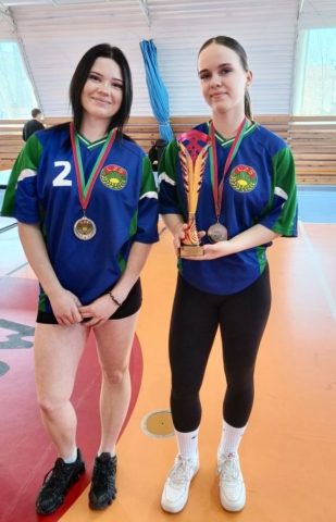 Dwie zawodniczki w strojach sportowych koloru zielonego, granatowego i czarnego z logo LZS oraz numerami startowymi na koszulkach stoją w hali sportowej. Obydwie mają na szyjach medale. Jedna z zawodniczek trzyma puchar.