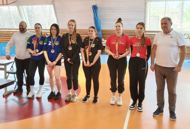 6 zawodniczek w strojach sportowych stoją w hali sportowej. Wszystkie mają medale na szyjach. 3 z nich trzyma też puchary. Widoczni też dwaj opiekunowie drużyn, stojący po prawej i lewej stronie zawodniczek.