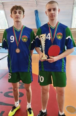 Dwóch zawodników w strojach sportowych koloru zielonego, granatowego i białego z białymi numerami startowymi na koszulkach i spodenkach oraz logo LZS na koszulkach stoi w hali sportowej. Obydwaj mają na szyjach medale, a jeden z nich trzyma w ręku puchar oraz paletkę do tenisa stołowego.