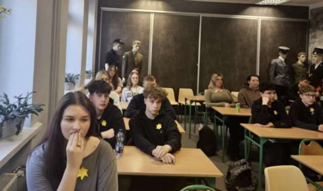 Młodzież szkolna siedzi w ławkach w sali konferencyjnej. Mają przypięte kotyliony w kształcie żonkili. W tle ściana koloru stalowego oraz manekiny w umundurowaniach.