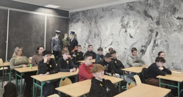 Młodzież szkolna siedzi w ławkach w sali konferencyjnej. Mają przypięte kotyliony w kształcie żonkili. W tle ściana koloru stalowego oraz manekiny w umundurowaniach.