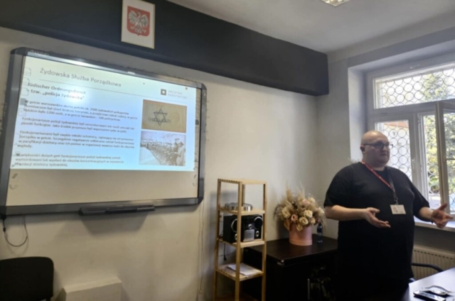 Mężczyzna noszący identyfikator na szyi stoi w sali konferencyjnej. W tle tablica multimedialna z wyświetloną prezentacją, slajd zatytułowany Żydowska Służba Porządkowa. W tle regał ze stojącym sprzętem muzycznym oraz komoda, na której stoi wazon z suszonymi kwiatami. Nad tablicą godło Polski.