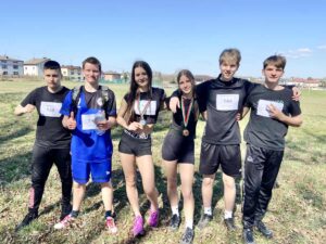 6 nastolatków, dwie dziewczyny i 4 chłopców w strojach sportowych stoją na zewnątrz. Dziewczyny mają na szyjach medale, 5 z osób ma numery startowe na koszulkach. Stoją obok siebie i pozują do zdjęcie wyrażając zadowolenie ze zdobytych trofeów. 