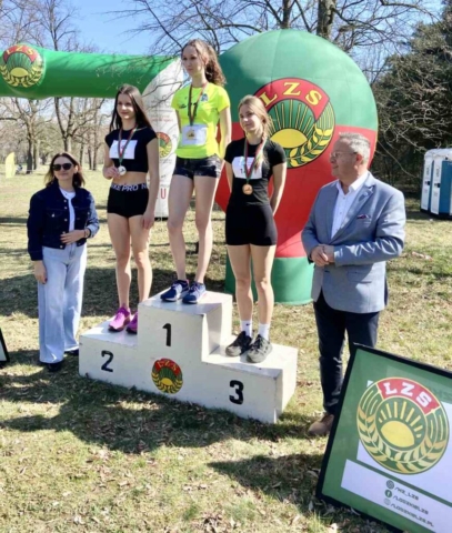 3 zawodniczki w strojach sportowych, stojące na podium na zewnątrz. Na koszulkach mają numery startowe, na szyjach medale zawieszone na szarfach zielono- czerwonych. W tle balon i meta z logo LZS. Po bokach podium dwie osoby dorosłe. Po prawej stronie rama z logo LZS oraz symbolami mediów społecznościowych LZS.
