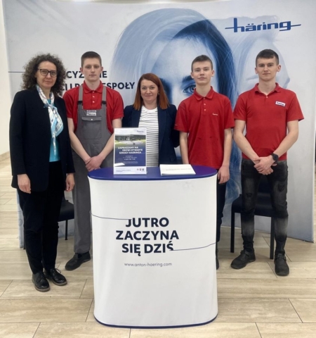 3 uczniów w strojach firmowych oraz dwóch opiekunów na stoisku promocyjnym firmy Haering na tle baneru reklamowego.