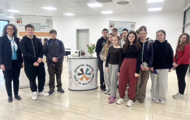 Po lewej stronie Dwóch uczniow i opiekun przy stoisku promocyjnym szkoły, ZS CKZ w Bujnach. W tle rollup z ofertą edukacyjną szkoły. Po prawej strone grupa młodzieży, uczestnicy dni otwartych.