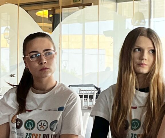 Dwie uczestniczki zagranicznych praktyk zawodowych w białych koszulkach z logo wyjazdu i napisem ZS CKZ Bujny siedzą w pomieszczeniu.