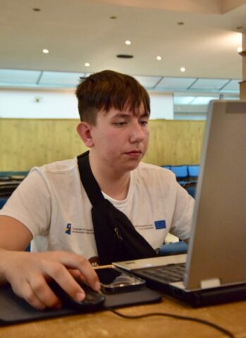 Uczestnik zagranicznych praktyk zawodowych, w białej koszulce wyjazdowych siedzi w pomieszczeniu przy biurku, na którym znajduje się laptop i pracuje nad czymś w skupieniu.