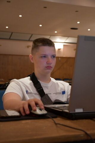 Uczestnik praktyki zagranicznej w białej koszulce wyjazdowej siedzi przy komputerze w pomieszczeniu.