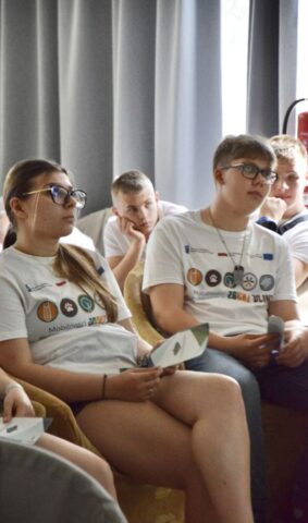 Młodziez uczestnicząca w praktykach zagranicznych siedzi w dużej sali konferencyjnej. Mają na sobie białe koszulki wyjazdowe.