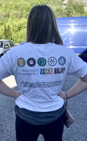 Uczestniczka praktyki zagranicznej stoi odwrócona tyłem na zewnątrz. Prezentuje koszulkę firmową koloru białego z logo i napisem Mobilności Europejskie z ZS CKZ Bujny.