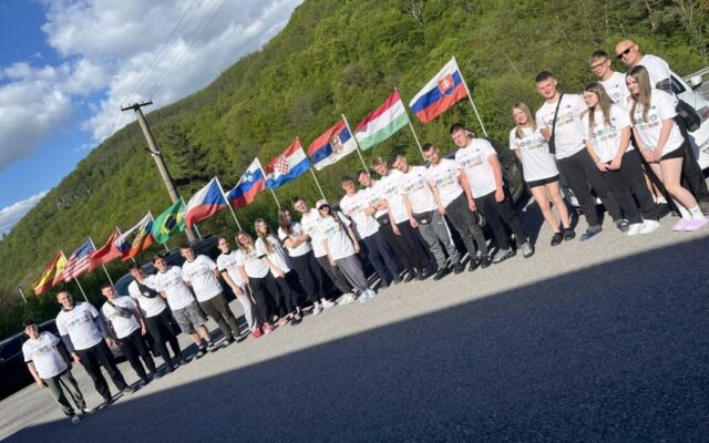 Grupa młodzieży wraz z opiekunami. Uczestnicy praktyki zagranicznej. Stoją na parkingu, w tle porośnięte drzewami wzgórza i rząd flag różnych krajów.