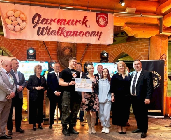 jarmark_wielkanocny01 Grupa młodzieży i osób dorosłych wewnątrz pomieszczenia hotelowego. Nad nimi baner w napisem Jarmark Wielkanocny, logo powiatu piotrkowskiego oraz po lewej stronie baneru koszyk z pisankami. Jedna z osób trzyma dyplom.,