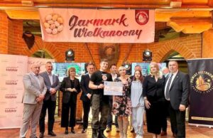 Grupa kilku osób stojących wewnątrz. Nad nimi baner z napisem Jarmark Wielkanocny, widoczny na nim też koszyk z pisankami i logo powiat piotrkowski. Pośrodku stoi uczeń i trzyma dyplom. 