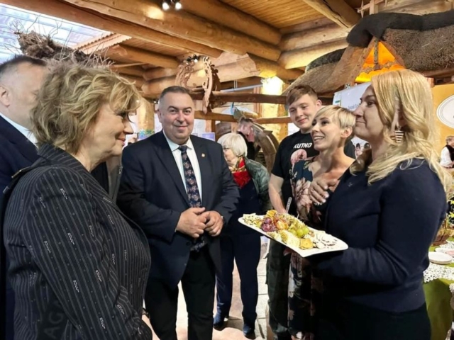 jarmark_wielkanocny05 Grupa osób stojących w pomieszczeniu o wystroju rustykalnym. Jedna z osób trzyma deskę serów przyozdobionych owocami.