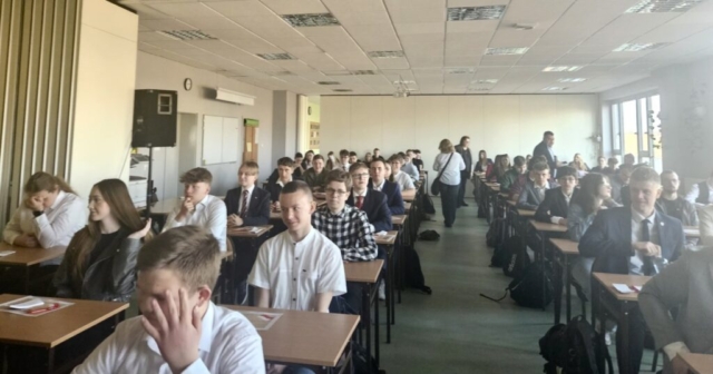 Duża sala konferencyjna, uczniowie siedzą w ławkach. W tle opiekunowie.
