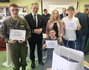 Laureaci konkursu, 3 uczniów trzymających w dłoniach dyplomy, wraz z opiekunem oraz patronem honorowym pozują do zdjęcia w klasie. W tle wystawa zdjęć. . Dwóch uczniów w mundurach klasy opw. W tle inni uczestnicy konkursu. 