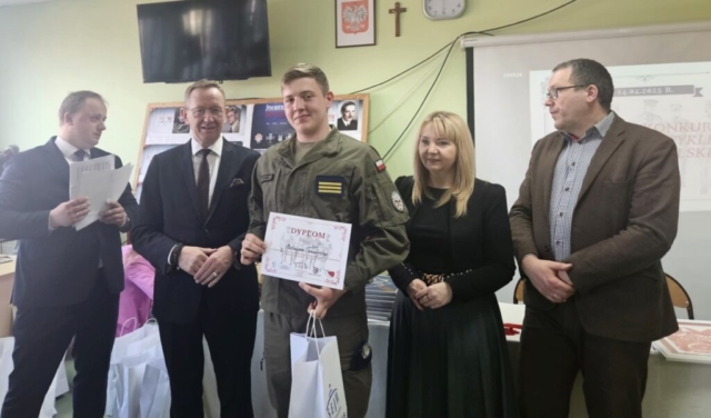 Klasa lekcyjna. Na pierwszym planie 4 organizatorów konkursu oraz uczeń w umundurowaniu klasy OPW, który trzyma w ręku dyplom. W tle ekran z wyświetlaną prezentacją oraz wystawa zdjęć.