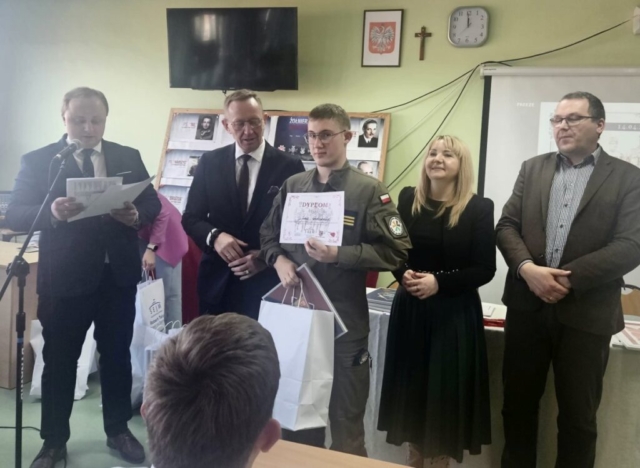 Klasa lekcyjna. Na pierwszym planie 4 organizatorów konkursu oraz uczeń w umundurowaniu klasy OPW, który trzyma w ręku dyplom. W tle ekran z wyświetlaną prezentacją oraz wystawa zdjęć.