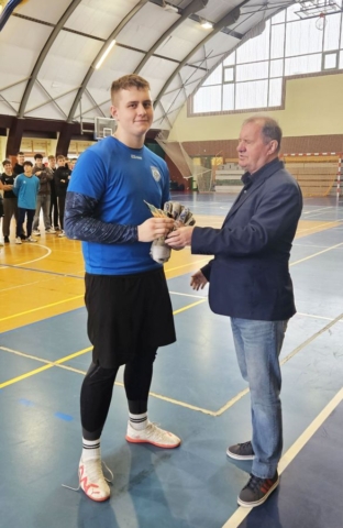 Zadownik w stroju sportowym czarno- niebieskim odbiera nagrodę i gratulacje od organizatora zawodów. Duża hala sportowa, w tle inni zawodnicy.