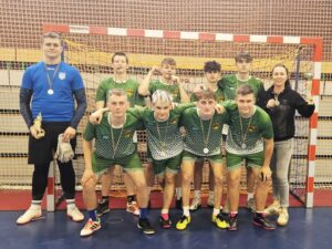Szkolna drużyna sportowa w strojach sportowych, 8 uczniów w strojach zielono- białych i jeden uczeń w stroju niebiesko- czarnym. Po prawej stronie również opiekun drużyny. Uczniowie mają na szyjach medale. Pozują w sali gimnastycznej na tle bramki. 