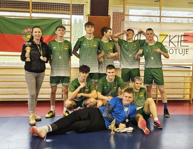 Drużyna sportowa wraz z opiekunem stoli w hali sportowej. Zawodnicy i opiekun mają na szyjach medale, opiekun dodatkowo trzyma zdobyte przez drużynę trofeum sportowe.