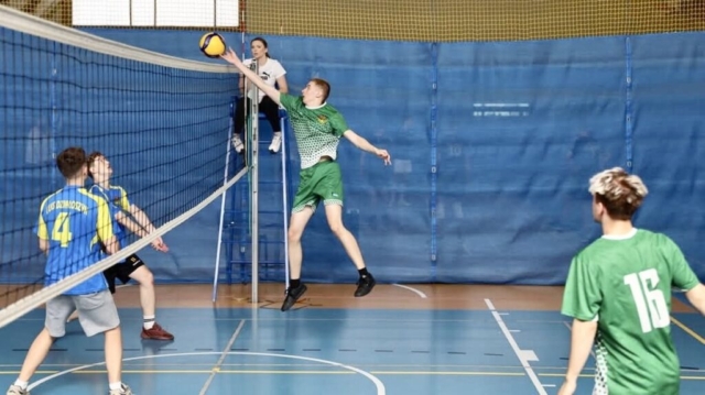 Kadr z meczu siatkówki rozgrywanego w hali sportowej. Po prawej stronie dwóch zawodników w strojach sportowych koloru zielonego i białego, widoczny jeden numer startowy 16. Po lewej stronie dwóch przeciwników, w strojach sportowych, niebieskie koszulki z żółtymi akcentami, szare i czarne spodenki, widoczny napis Działoszyn i numery startowe. Pośrodku na stanowisku sędziowskim kobieta w białej koszulce i czarnych spodniach, która sędziuje mecz.