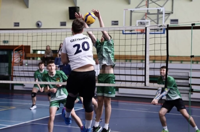 Kadr z meczu piłki siatkowej. Na pierwszym planie zawodnik z numerem startowym 20 i nazwiskiem Grzybowski na białej koszulce, ma też czarne spodnie i ochraniacze. Po stronie przeciwników zawodnicy w strojach zielonym i białym bronią piłki. W tle trybuny sportowe i 2 kibiców.