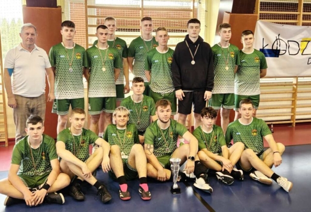 Drużyna sportowa z organizatorem. Zawodnicy mają na szyjach medale, górny rząd stoi, dolny rząd siedzi, przed nimi stoi wygrany puchar. W tle drabinki sportowe oraz po prawej stronie część baneru z napisem Łódź.